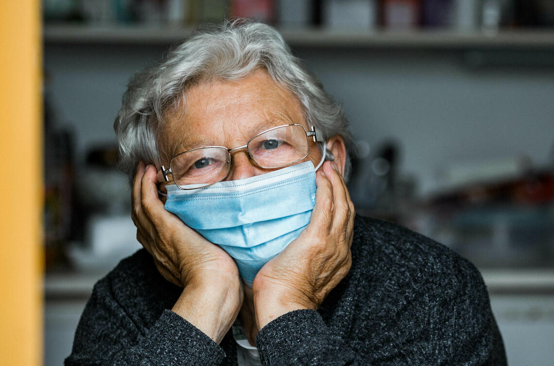 Older woman wearing a medical mask, looking out a window